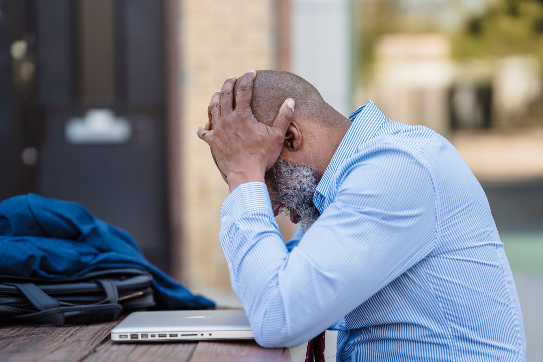 Side view of pensive middle aged African American male entrepreneur wearing blue shirt grabbing head with hands while si