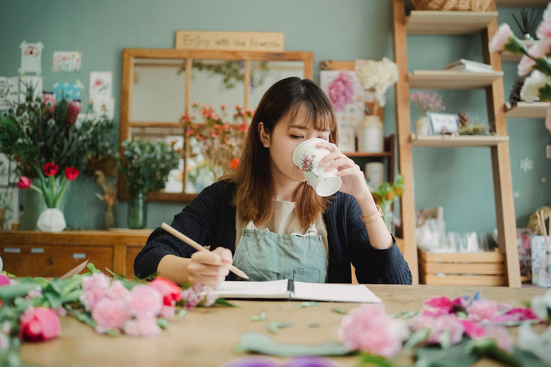 Concentrated Asian female florist drinking hot beverage and sitting at table with flowers while taking notes in notebook