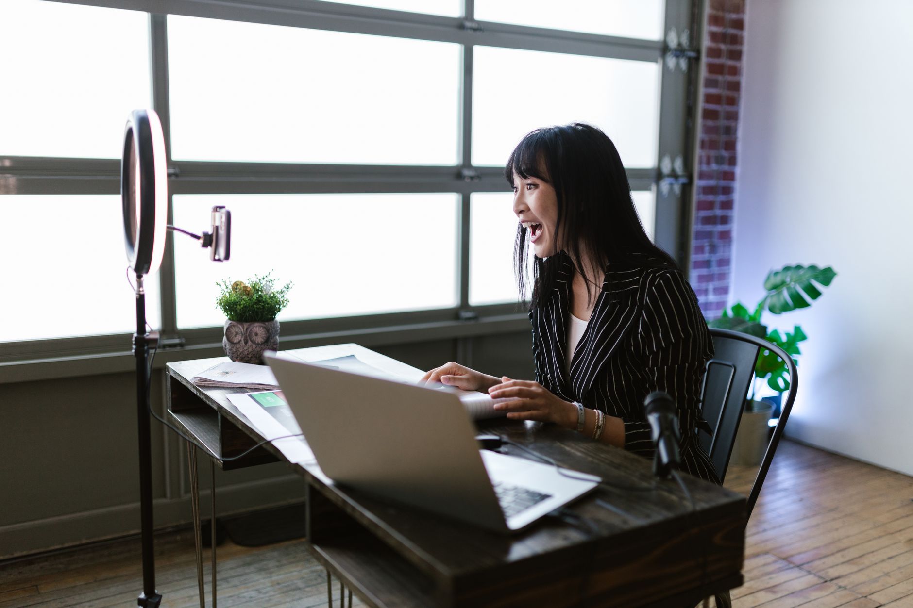 Asian woman in office setting recording content with laptop and ring light, showcasing elegant business setup.
