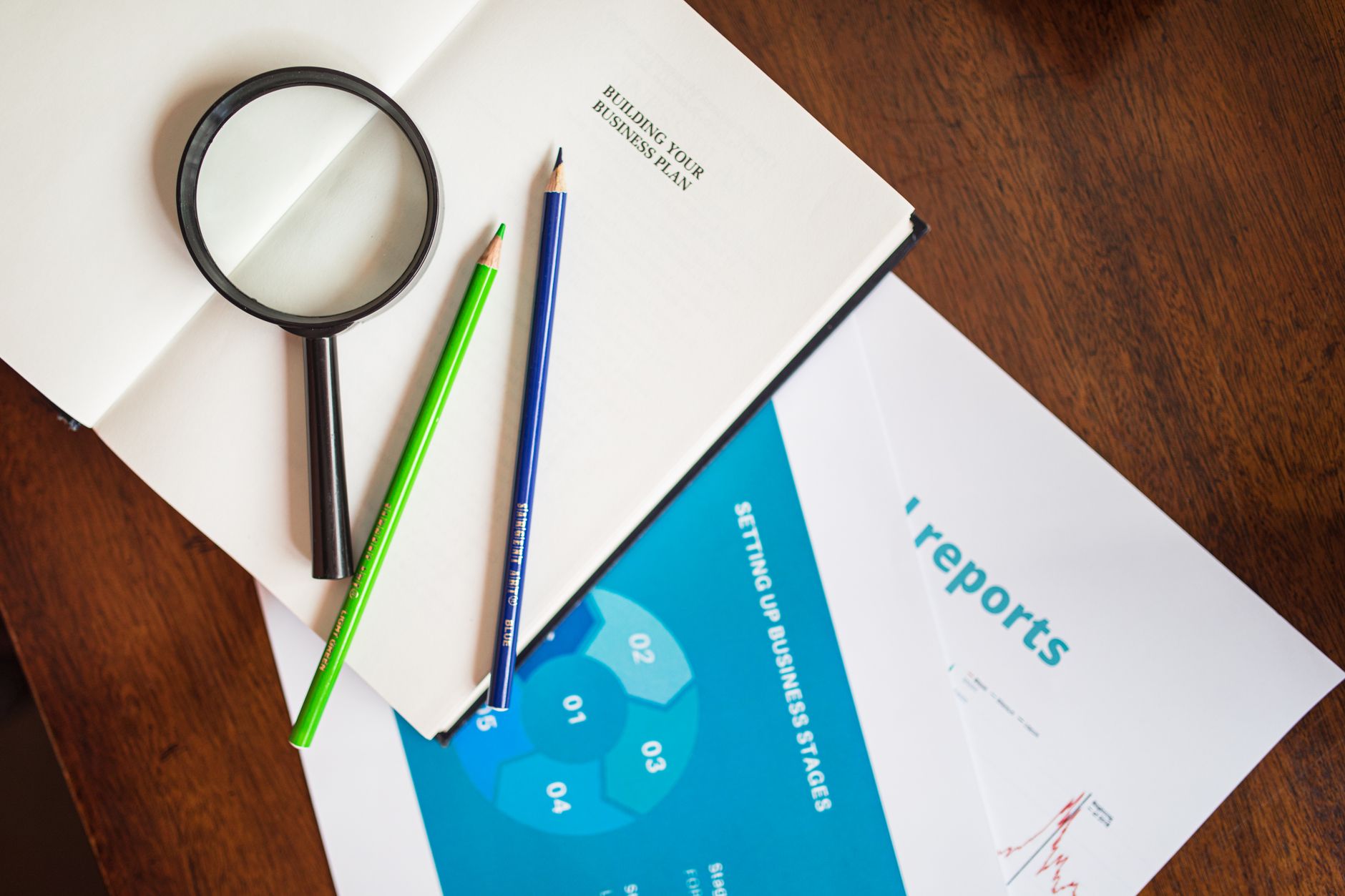 A top view of a magnifying glass, colored pencils, and business reports on a wooden table.