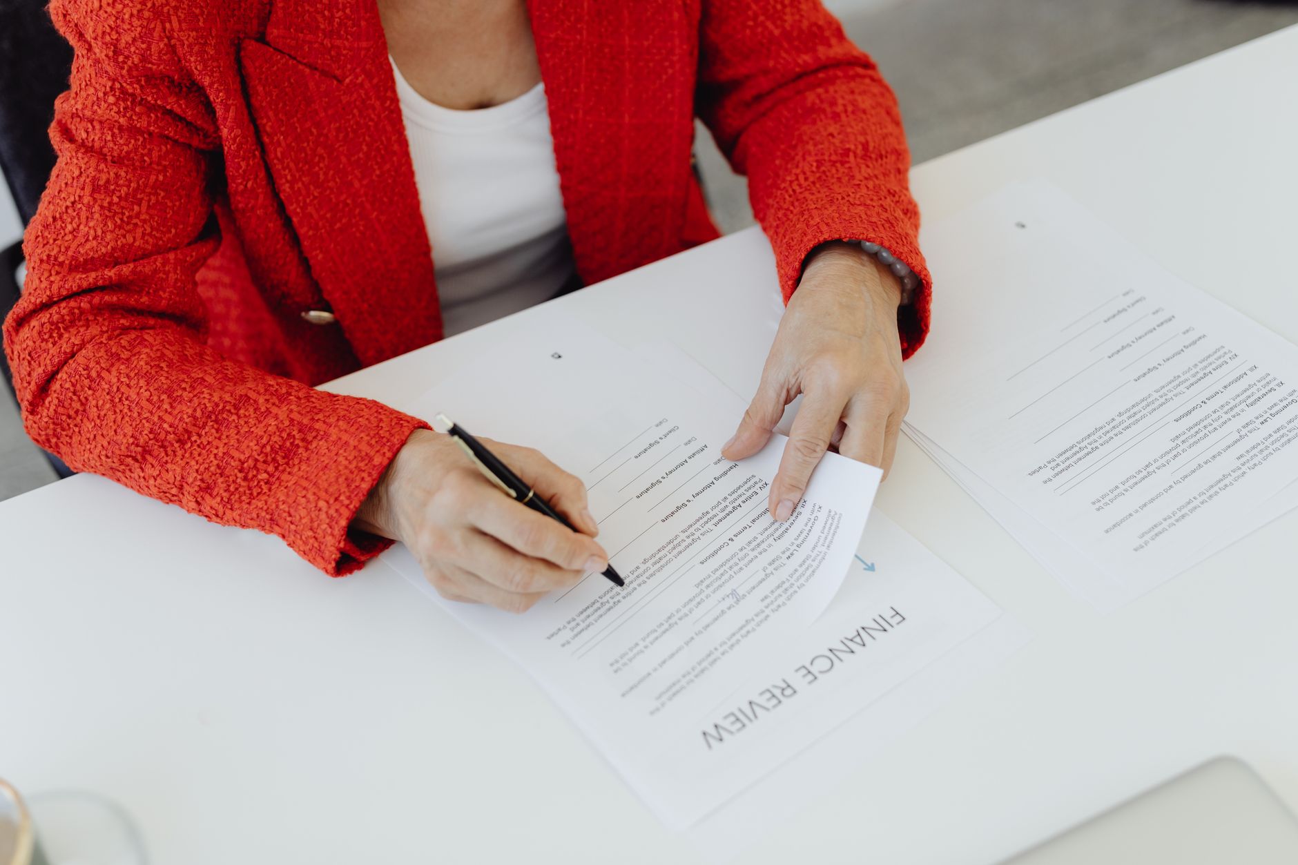 Close-up of a businesswoman in a red blazer signing finance documents at an office desk.